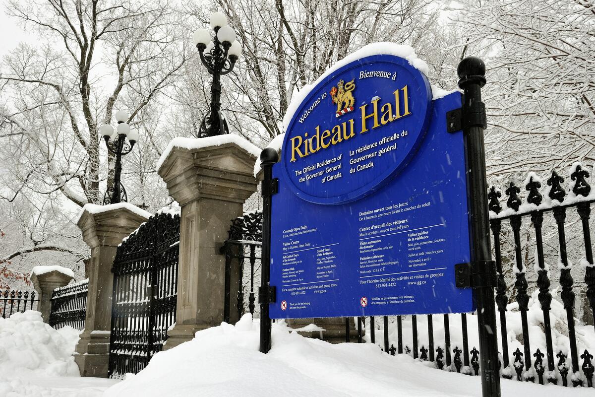 Snow-covered sign for Rideau Hall, the official residence of Canada's Governor General. Set against an ornate iron fence and winter trees.