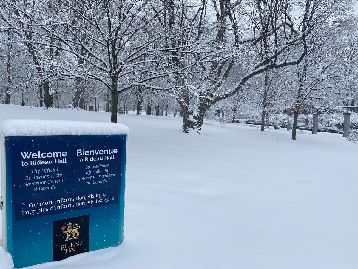 Snow-covered landscape with bare trees, a sign reads "Welcome to Rideau Hall, The Official Residence of the Governor General of Canada." Serene winter scene.