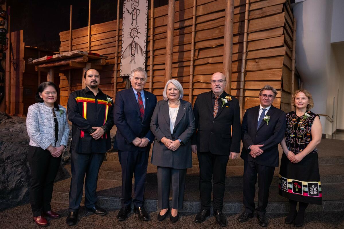 Governor General Mary Simon and Mr. Whit Fraser are standing in the centre of a line of 5 other people, in front of a wooden wall. Everyone is smiling at the camera.