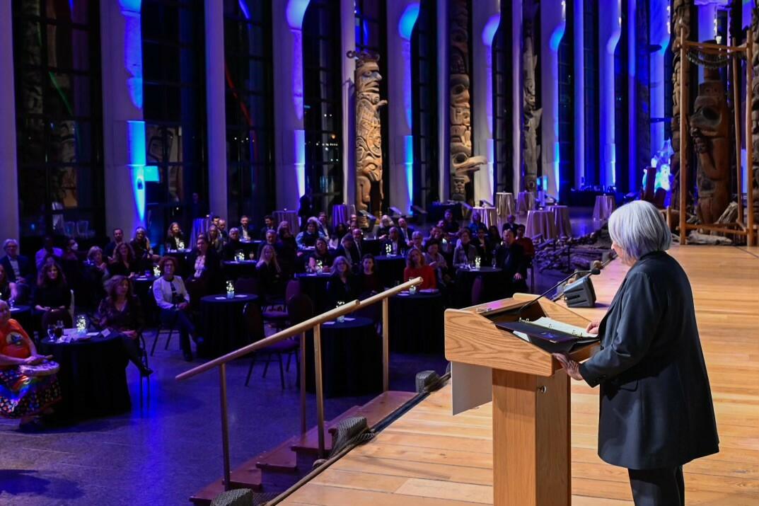 Governor General Mary Simon is seen from the left side, speaking at a podium, in front of a crowd of people sitting at round tables. Purple and lite blue lights are projected on tall pillars behind the audience.