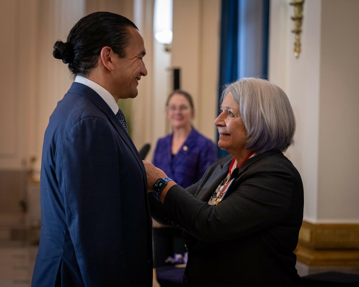 Governor General Simon is smiling to Premier Wab Kinew while pining a Coronation Medal on his vest.