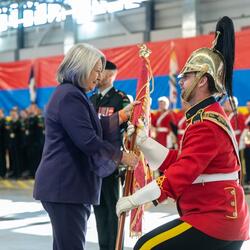 La gouverneure générale Mary Simon porte un costume violet et sourit à un membre des Forces armées canadiennes alors qu'elle lui remet le guidon. Le membre des Forces armées canadiennes porte un uniforme rouge, des gants blancs et un casque. 