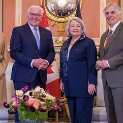 Governor General Simon Stands next to His Excellency Frank-Walter Steinmeier, President of the Federal Republic of Germany, Mr. Whit Fraser and Ms. Elke Büdenbende. They are smiling at the camera. Behind them is the flag of Canada and the flag of Germany.