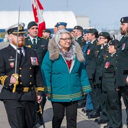 Governor General Simon walking outside. A man in a military uniform is walking beside her, to her left. To her right, there is a row of individuals in military uniforms. 