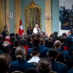 La gouverneure générale Simon parle dans un microphone devant un grand nombre de personnes. Les murs de la salle sont bleus. Un grand miroir orné d'or se trouve derrière Son Excellence.