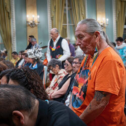 A silver haired man in an orange shirt stands among seated and standing peers.