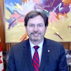 Screenshot of the Administrator smiling at the camera. There is a Canadian flag to his left at a brightly coloured painting behind him. 