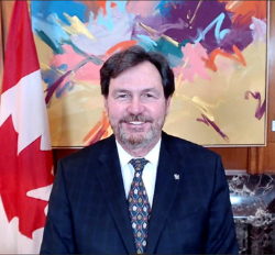 Capture d'écran de l'administrateur souriant à la caméra. Il y a un drapeau canadien à sa gauche et une peinture aux couleurs vives derrière lui. 