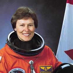 L'astronaute canadienne Roberta Bondar dans un uniforme d'astronaute orange avec un casque sur une table. Un drapeau du Canada est à l'arrière-plan.