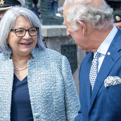 Governor General Mary Simon, wearing a blue vest, is smiling and talking to His Majesty the King Charles III, wearing a navy blue suit and a blue tie.