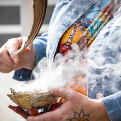A person holding a bowl with rising smoke, using a feather to spread it. 