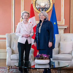 Governor General Simon Stands next to Her Excellency Ursula von der Leyen, President of the European Commission. They are smiling at the camera. Behind them is the flag of Canada and the flag of Europe.