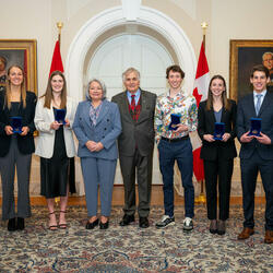 A group photo featuring Governor General Simon, Mr. Whit Fraser and seven recipients of the Governor General’s Academic All-Canadian Commendation. The recipients are each holding a blue box with a medallion inside of it. Behind them: two Canadian flags.