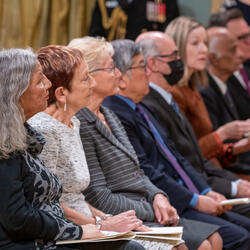 Four women and four men sitting on chairs. Several of them hold booklets. They are looking ahead.