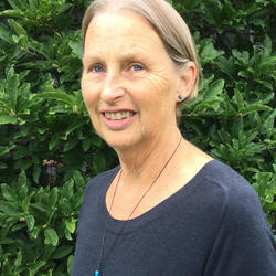 Member of the Order of Canada - Lucille Harper. She is wearing a navy blue top with a necklace. She is standing outside. There are many trees and bushes behind her.