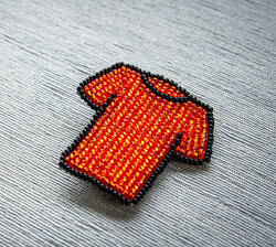 A pin of a mini orange t-shirt made of beads with black outline on a grey background.