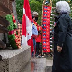 La gouverneure générale dépose une couronne au pied du Monument national aux anciens combattants autochtones. On voit des plumes à la gauche de la photo. On voit des arbres ainsi que des gens portant des masques derrière elle. 