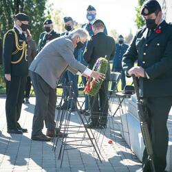 Mr. Whit Grant Fraser is laying a wreath in front of a stone memorial. Several members of the Canadian Armed Forces are standing around him, wearing masks. 