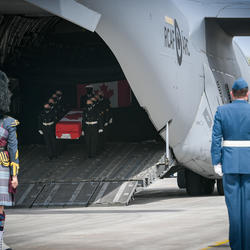 At the forefront, we see the back of Canadian Armed Forces(CAF) members, including a bagpiper. In the background, we see a coffin, carried by CAF members, being taken off a plane. 