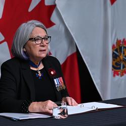 La gouverneure générale Mary Simon est assise à une table. Devant elle il y a un cahier ouvert et derrière elle on peut voir le drapeau canadien et le drapeau des Forces armées canadiennes.