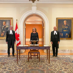 La gouverneure générale est debout, devant une porte ouverte. Une table et une chaise de bois est devant elle. Deux hommes sont à sa gauche. Un homme et une femme sont à sa droite. Tous sont alignés.