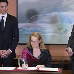 The Governor General signs the oath book. The Prime Minister and the Clerk of the Privy Councillor are on each side of her. 