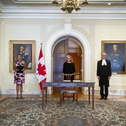 Five people standing in front of a pastel yellow wall. A table is in front of the person standing in the middle. A Canadian flag is placed to her right.