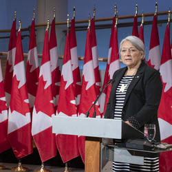 Le premier ministre Justin Trudeau et la gouverneure générale désignée Mary May May Simon se tiennent chacun face à un podium devant plusieurs drapeaux du Canada derrière eux. Mary Simon parle. Justin Trudeau la regarde.