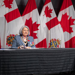 Governor General Mary Simon is sitting at a long table with General Wayne Eyre. They are both facing the camera. Behind them are Canadian and Canadian Armed Forces flags.