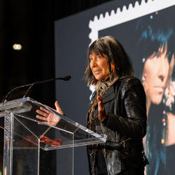 Buffy Sainte-Marie speaks at a podium. A photo of her new commemorative stamp is visible behind her.