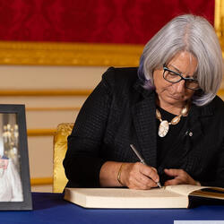 Governor General Mary Simon is sitting at a table and writing in a book of condolences. There is a portrait of Her Majesty The Queen on the table. Text in the lower right corner of the image reads, “Credit: David Parry/PA Media Assignments.”