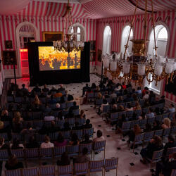 Une grande foule de personnes est assise dans la salle de la tente de Rideau Hall et regarde un film sur grand écran