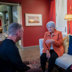 A group of people gathered in a warmly lit room, seated on patterned sofas. Governor General Mary Simon is engaged in conversation. A few people stand in the background by a table with tea and coffee.