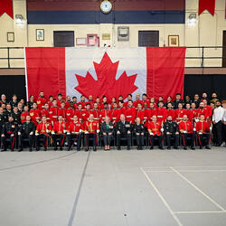 Governor General Mary Simon and a large group of military personnel and civilians posing for a group photo in front of a Canadian flag. The group includes soldiers seated and standing in rows in a large gymnasium.