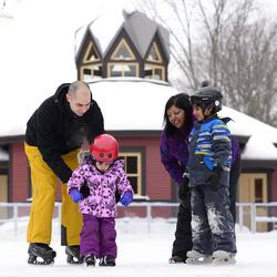 Une famille de 4 (2 parents and 2 jeunes enfant) qui patinent.  Le pavillon d’hiver est en arrière-plan.