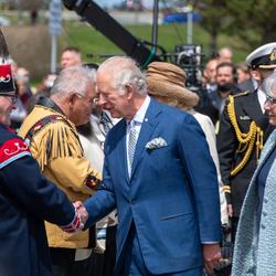 His Royal Highness The Prince of Wales shakes the hand of an Indigenous leader as he walks in front of a row of people. Governor General Simon walks to his left.