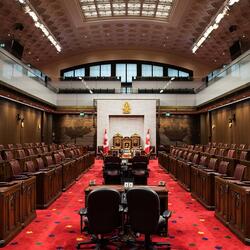 Empty Senate of Canada chamber. Several dark desks down the middle aisle. Dark brown leather chairs in seating areas on either side. A large throne at the front of the room. A glass-balcony gallery looks down on the space. 