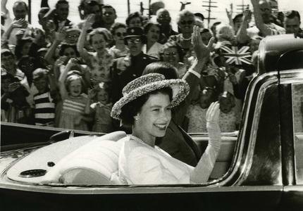 Photo en noir et blanc d’une jeune reine Elizabeth II. Elle salue la foule depuis le siège arrière d’une voiture décapotable. Elle porte un chapeau, un collier de perles et des gants.