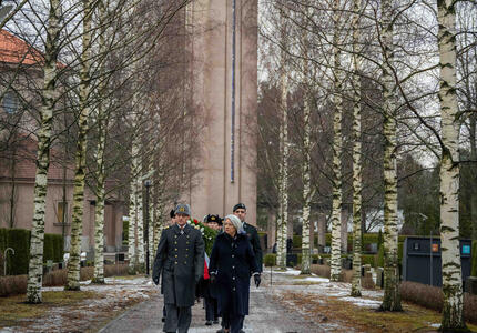 La gouverneure générale Mary Simon marche sur un chemin de gravier dans un cimetière. Un homme en uniforme marche à côté d’elle et un groupe de personnes marche derrière eux. Derrière le groupe se trouve une grande tour d’horloge en pierre.