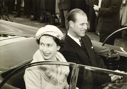 The Queen and the Duke of Edinburgh smile at the crowd while riding in an open-air vehicle. The photo is black and white.