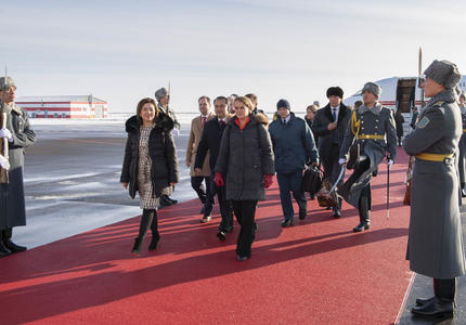 The Governor General walks on a red carpet which leads to the stairs of an airplane in the background.  She is surrounded by officials.  Armed guards line the red carpet.