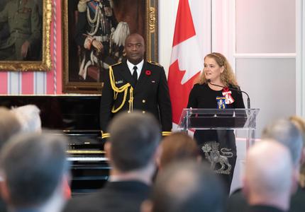 The Governor General stand at a podium and delivers a speech.  Behind her is her Aide-de-Camp in a Navy uniform as well as the Canadian flag.  