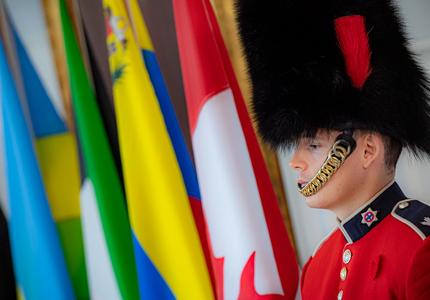 A member of the Governor General’s Foot Guards is standing in front of flags. 