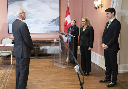 Michael Wernick, clerk of the Privy Council, stands in front of Governor General Julie Payette and Prime Minister Justin Trudeau.   In the corner of the room stands the MC behind a podium. 