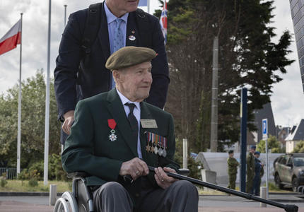 A war veteran in a wheelchair, wearing a beret and war medals, is pushed by a man. Several flags are in the background.