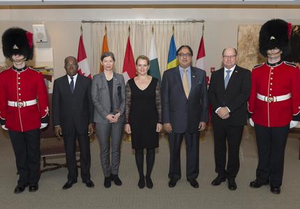 A groupe photo of the four news heads of mission with the GOvernor General.  They are flanked by ceremonial guards and stand in front of a series of flags representing each country. 