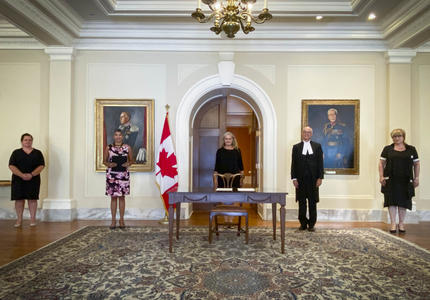 Cinq personnes sont alignées contre un mur jaune pâle. Une table est devant la personne au centre. Un drapeau du Canada se trouve à sa droite.