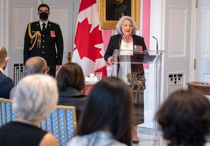 La gouverneure générale Mary May Simon, debout derrière un lutrin en verre, s’adresse au public rassemblé dans la salle de la tente de Rideau Hall. Un imposant drapeau canadien décore le fond de la pièce.