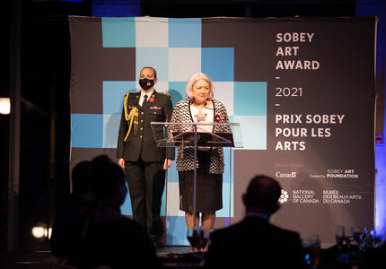 The Governor General stands at a podium at the 2021 Sobey Art Award Ceremony. A member of the military stands behind her.