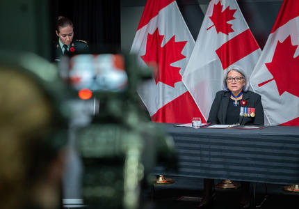 Une caméra enregistre la gouverneure générale Mary Simon. On la voit sur l'écran LCD de la caméra. Elle est également à l'arrière-plan de la photo. Derrière elle, on aperçoit des drapeaux canadiens.